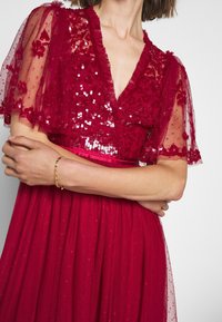 Femme portant une robe rouge profond avec un corsage floral à sequins et des manches transparentes et flottantes, bras croisés montrant un bracelet chaîne en or.