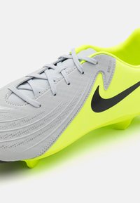 Botas de fútbol sintéticas grises con una superficie texturizada, logo de Nike en negro y acentos en amarillo brillante. Presenta cordones grises y tacos moldeados.