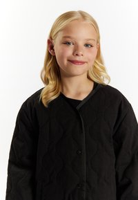Sort quiltet jakke med et struktureret mønster, der har en rund halsudskæring og knaplukning, præsenteret mod en hvid baggrund.