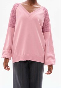 Personne portant un pull rose ample à col en V avec des détails en dentelle sur les épaules et un pantalon à fines rayures sombres sur un fond blanc.