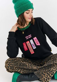 Černá mikina s barevnými štítky "LÁSKA", "RYTMI", "RADOST" a "RIZIKO", nošená přes kalhoty s leopardím potiskem a doplněná o zelenou čepici.