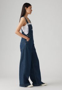 Blå denim overaller med vide ben, justerbare stropper, sidelommer og en liten frontlomme, iført over en hvit topp.