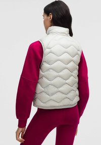 Jasnoszara pikowana kamizelka z wysokim kołnierzem, zestawiona z długim różowym topem. Charakteryzuje się gładką, błyszczącą fakturą.