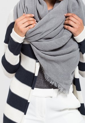 Écharpe grise en matière tissée douce, portée élégamment autour du cou avec des bords à franges. Portée avec un pull à rayures et un pantalon blanc.