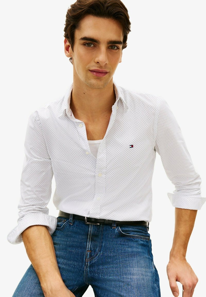 Jeune homme aux manches retroussées portant une chemise blanche à motifs boutonnée par-dessus un sous-vêtement blanc et un jean bleu avec une ceinture noire.