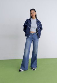 Casaco corta-vento azul-marinho com detalhes brancos, top cinza curto e jeans azuis flare. Ténis em preto e branco completam o visual. Chão verde.