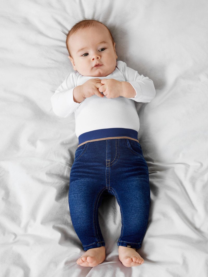 Bebé vistiendo una camisa blanca de manga larga y pantalones estilo mezclilla azul con una cinturilla el�ástica, tumbado sobre una manta de color claro.