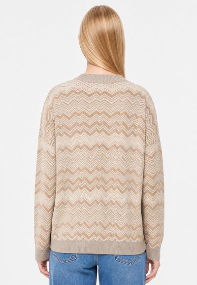 Femme aux longs cheveux blonds portant un pull beige et blanc à motif zigzag et un jean bleu, vue de dos sur un fond uni.