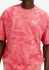 Růžové tričko s krátkým rukávem ve stylu tie-dye vyrobené z měkké tkaniny. Obsahuje bílé logo na hrudi a má volný střih s žebrovaným výstřihem.