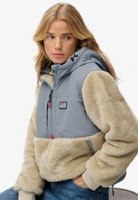 Puffer-Weste mit einer hellgrauen Schale und cremefarbenem, flauschigem Stoff. Verfügt über einen Reißverschluss vorne, eine Kapuze und ein buntes Logo-Patch auf der Brust.