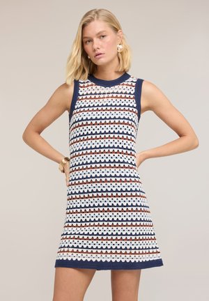 Blondynka w bezrękawnej, sięgającej do kolan sukience na szydełku w poziome paski w kolorach granatowym, białym i rdzawego, pozująca z rękami na biodrach.