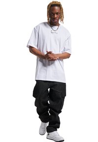 White cotton T-shirt with short sleeves and a printed logo. Dark cargo pants with side pockets. White sneakers. Model stands with hands clasped.