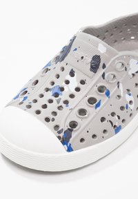 Zapato gris ventilado con un patrón de salpicaduras en azul y negro, con puntera de goma y suela con diseño. Su diseño ligero incluye múltiples perforaciones.