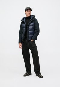 Jonge man die staat met een zwarte pet, een marineblauwe pufferjack, een zwarte coltrui, losse zwarte jeans en zwarte leren schoenen op een witte achtergrond.