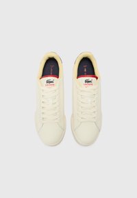 Cream-colored sneakers with a smooth texture, featuring round toes, lace-up design, and a red and navy logo tag on the tongue.