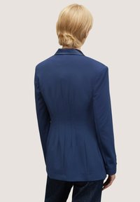 Blazer blu navy su misura con un design strutturato, parte anteriore monopetto e dettaglio plissettato sul retro, realizzato in tessuto liscio.
