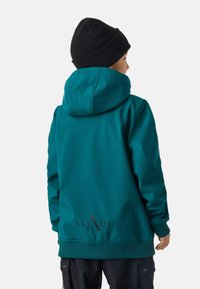 Felpa con cappuccio color verde pavone, vestibilità comoda, polsini e orlo a costine, con logo nero nella parte bassa della schiena. Indossata con un berretto lavorato a maglia nero.