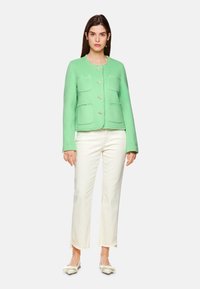 Grüne strukturierte Jacke mit vier vorderen Taschen, Knopfverschluss und rundem Halsausschnitt, kombiniert mit cremefarbenen, weit geschnittenen Hosen und hellen Schuhen.