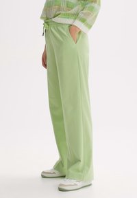 Světlé zelené široké kalhoty s pasem na stažení, bočními kapsami a hladkým povrchem, doplněné bílými a béžovými teniskami.