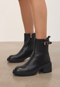 Botas de tobillo de cuero negro con cremallera lateral y hebilla decorativa, llevadas en los pies contra un fondo de pared y suelo neutro.