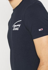 T-shirt uomo a maniche corte blu navy con logo bianco "Tommy Jeans" sul petto e piccolo emblema della bandiera rossa, bianca e blu sulla manica.