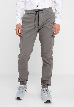 Mężczyzna noszący szare dresowe spodnie bojówki z czarnym sznurkiem i elastycznymi mankietami, w połączeniu z jasnoszarymi butami Nike i białą koszulą w wzory.