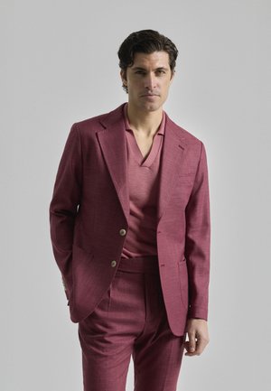 Traje marrón oscuro hecho de tejido texturizado, con una chaqueta de doble botonadura y una camisa de cuello en v, combinado con pantalones a medida.