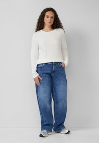 Weißes, geripptes Langarmshirt, getragen mit lässigen blauen Jeans. Die Jeans verfügen über ein klassisches Five-Pocket-Design und eine ausgewaschene Textur. Silberne Sneaker.