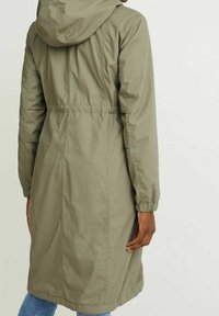 Olivgrön lång jacka med huva, åtdragen midja, elastiska manschetter och ett slätt, texturerat tyg. Bakvy som visar sömmarna och formen.