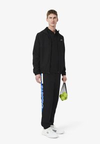 Zwarte zip-jack over een zwart poloshirt, met zwarte broek met blauwe "LACOSTE" tekst. Witte sneakers en een nettas met groene tennisballen.