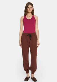 Kobieta stojąca twarzą do przodu, ubrana w bezrękawnik w kolorze magenty, brązowe spodnie dresowe z troczkiem i ciemnobrązowe mokasyny na białym tle.