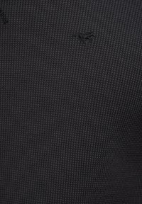 Melnas teksturētas kreklas audums ar vafelīša rakstu; augšējā kreisajā stūrī redzama neliela izšūta logo.