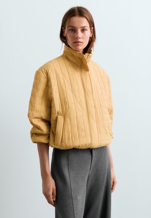 Une jeune femme porte une veste courte matelassée jaune à col montant et un pantalon gris ample, debout devant un fond uni.
