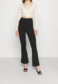 Beiges, langärmliges kurz geschnittenes Oberteil mit V-Ausschnitt, kombiniert mit hoch taillierter schwarzer Schlaghose, getragen mit schwarzen offenen Sandaletten.