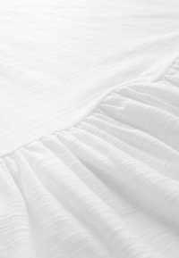 Bílá látka s jemným, mírně texturovaným povrchem. Jeden okraj zdobí nařasené volány, což vytváří jemný zavěšený vzhled.
