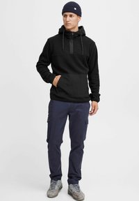 Schwarzer Kapuzenpullover mit halbem Reißverschluss, strukturiertem Frontpanel und großer Kängurutasche; kombiniert mit marineblauen Cargo-Hosen und grauen Turnschuhen.