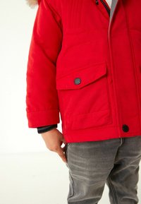 Röd isolerad jacka med dragkedja och knäppning, två framfickor med svarta knappar, i kombination med grå denimbyxor.