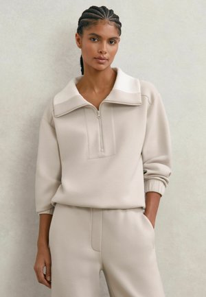 Beige quarter-zip trui met een opgevouwen kraag, gestructureerd voorpand en geribbelde manchetten, gecombineerd met bijpassende taps toelopende broek. Gladde textuur.