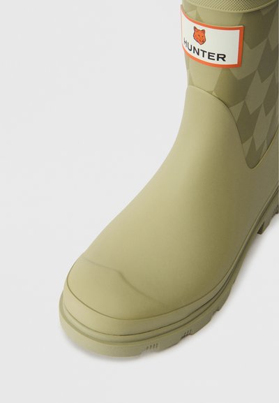 Olivno zeleni gumijasti škorenjček za dež z teksturirano, diamantno vzorčasto površino, zaobljenim nosom in oznako logotipa v oranžni in beli barvi blizu vrha.