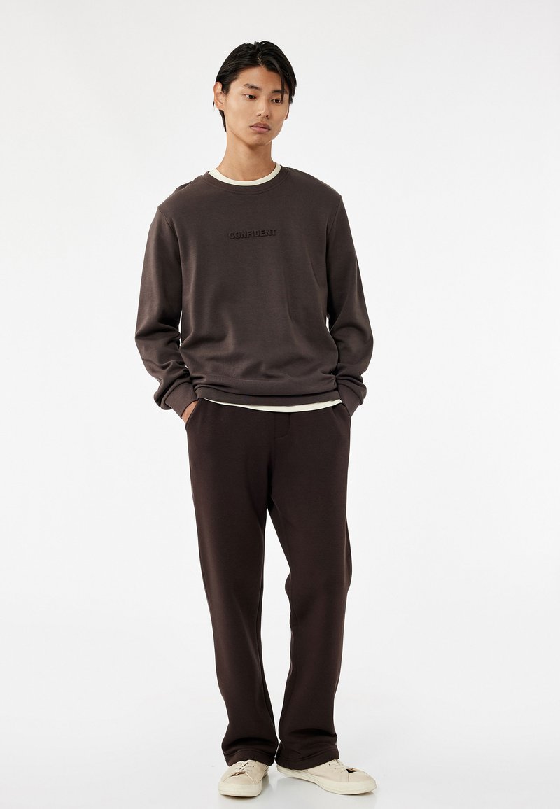 Brun sweatshirt med teksten "CONFIDENT", parret med matchende brune bukser. Materialet ser blødt ud; afslappet pasform; båret med hvide sneakers.
