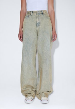 Osoba nosząca jasne, szerokie spodnie dżinsowe z wysokim stanem, białe trampki oraz biały top bez rękawów, stojąca na białym tle.