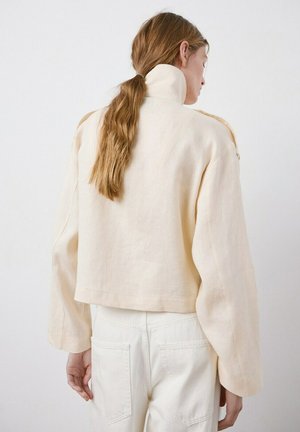 Mujer con cabello largo y castaño recogido en una coleta baja, vestida con una chaqueta corta de cuello alto en color crema y pantalones blancos, de pie frente a un fondo liso.