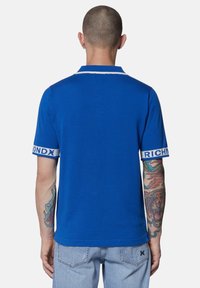 Polo blu royal con colletto bianco e testo "RICHMOND" a contrasto sulle maniche. Design aderente, texture liscia, mostrato da dietro.