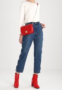 Kobieta w białej koszuli z długim rękawem, w wysokich niebieskich dżinsach z bocznymi guzikami, w czerwonych lakierowanych botkach na kostkę oraz z czerwoną torbą na ramię.