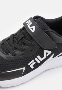 Chaussure de sport noire avec une tige en mesh, fermeture à lacets et velcro, marquage blanc et une semelle en caoutchouc blanche avec un design géométrique.