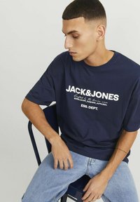 Camiseta de gran tamaño en azul marino con texto blanco que dice "JACK&JONES" y detalles gráficos. Llevada con jeans de color azul claro, sentado en una silla.