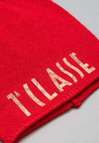 Cappellino rosso lavorato a maglia con scritta "TILASSIE" in lettere con texture effetto sughero. Presenta un orlo a coste e una costruzione senza cuciture. Texture morbida.