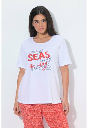 Kobieta z długimi ciemnymi włosami, ubraną w białą koszulkę z czerwonym nadrukiem i czerwone spodnie w wzory, stojąca na białym tle.