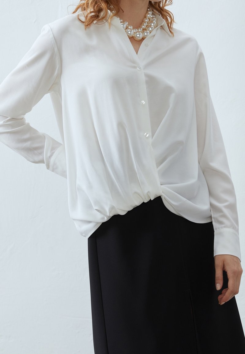 Camicia bianca a maniche lunghe con bottone frontale, caratterizzata da un nodo arricciato in vita, abbinata a una gonna nera. Tessuto morbido e leggero.