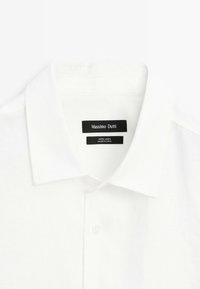 Biała lniana koszula z klasycznym kołnierzykiem i pojedynczym zapięciem na guzik. Metka z napisem "Massimo Dutti" oraz informacjami o materiale. Gładka struktura.